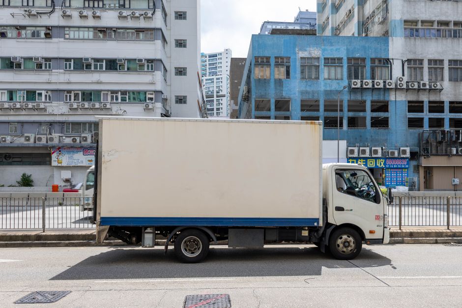 A small white moving van with a cargo box parked on a narrow residential street, surrounded by multi-storey brick and concrete buildings with various window styles and rooftop structures. The van is p