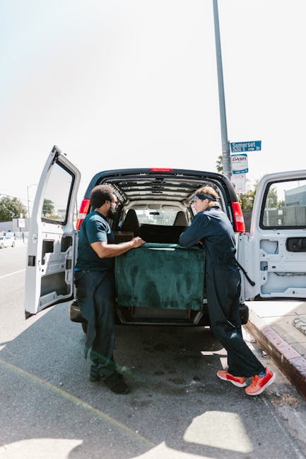 Two movers from [COMPANY_NAME] are loading a large piece of furniture wrapped in green moving blankets into the rear of a white commercial van during a home relocation in an outdoor urban setting. The